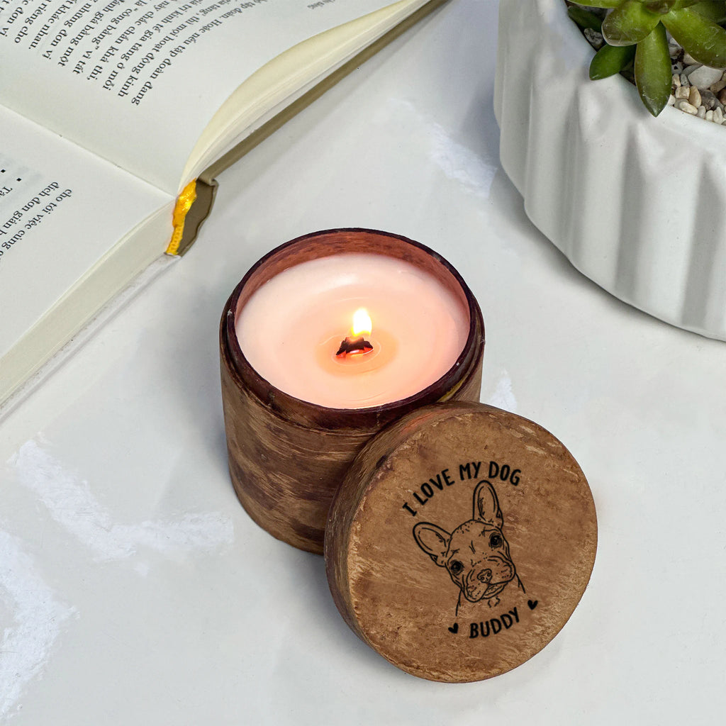Candle in a wooden container with 'I love my dog' engraving on a white surface with an open book and plant.