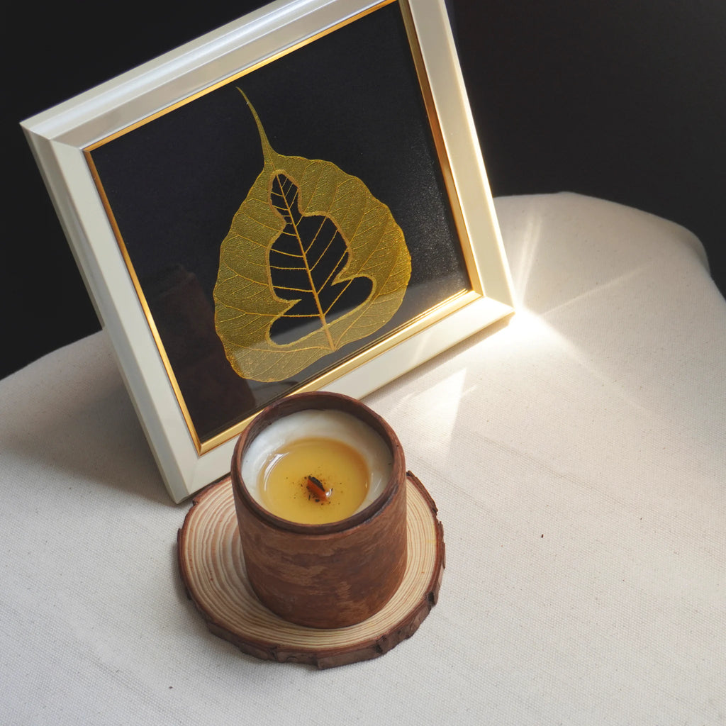 Candle in a wooden holder with a framed leaf art piece on a white surface