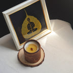 Candle in a wooden holder with a framed leaf art piece on a white surface