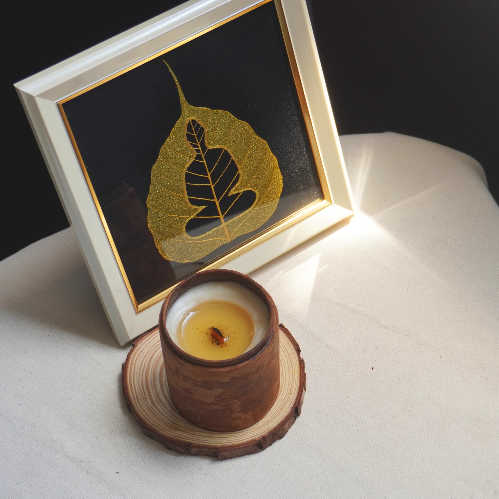 Candle in a wooden holder with a framed leaf art piece on a white surface