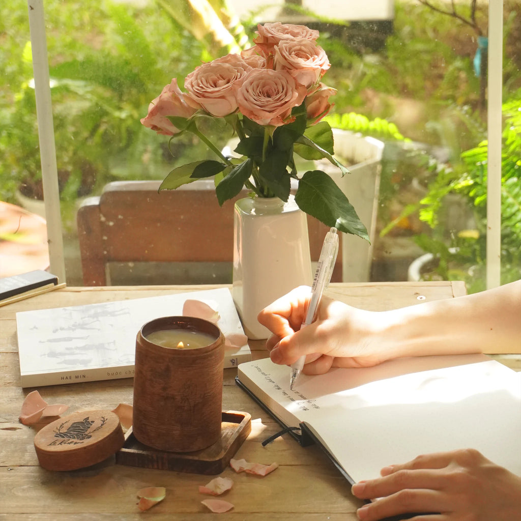 Person writing in a notebook with a candle, flowers, and book on a wooden table.