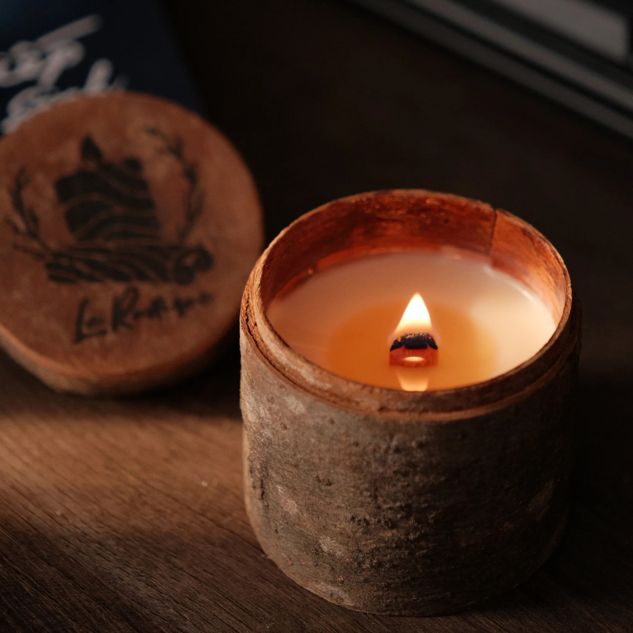 Candle in a rustic container with a wooden lid featuring branding on a dark background