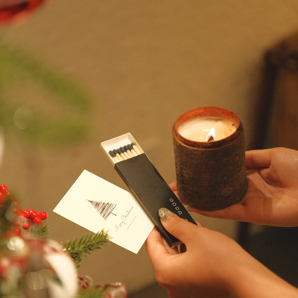 Person holding a lit candle and a lighter with matches, surrounded by festive decorations.