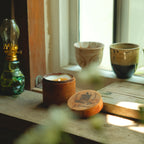 natural candle in a cinnamon wooden holder with a green oil lamp and ceramic cups on a windowsill.