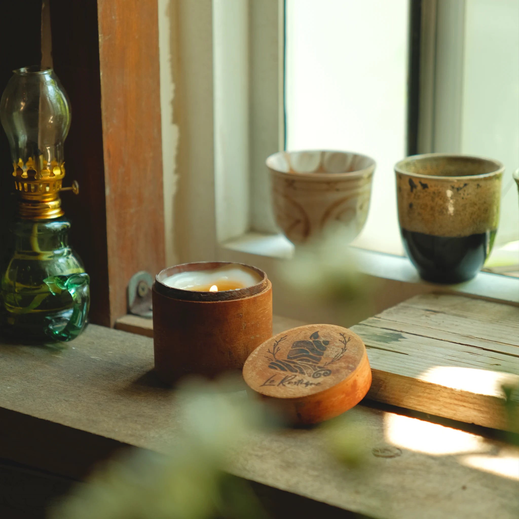 natural candle in a cinnamon wooden holder with a green oil lamp and ceramic cups on a windowsill.